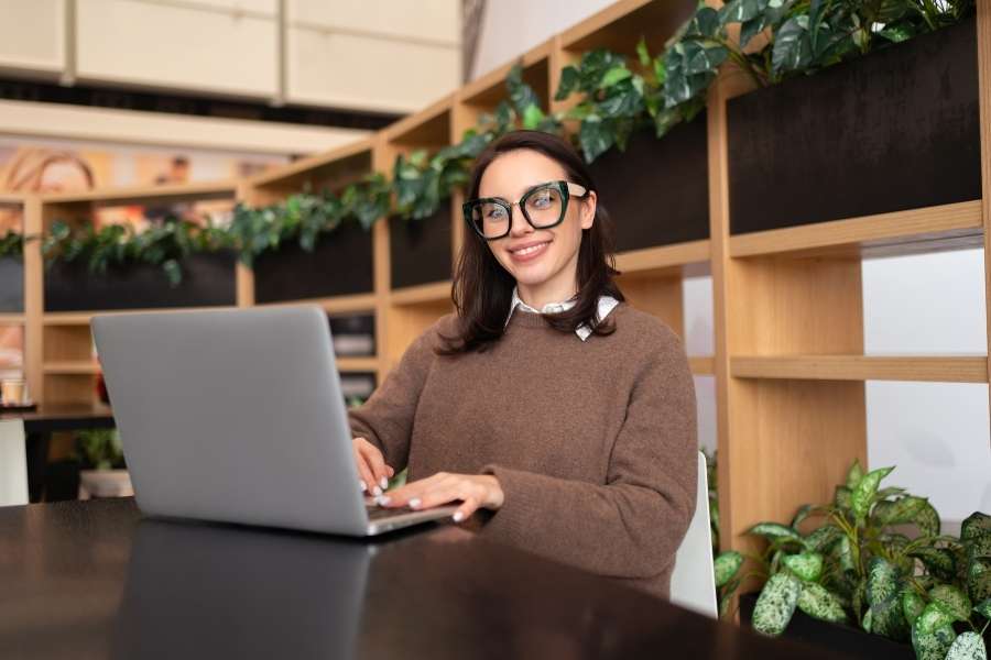 Une jeune femme souriante avec des lunettes utilise son laptop pour la première fois dans un espace de travail verdoyant.