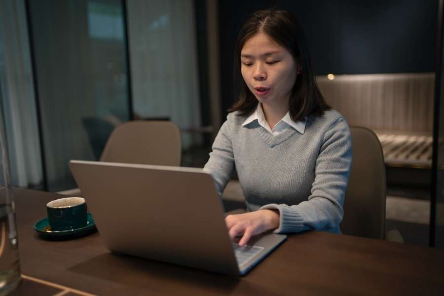 Une femme concentrée, assise à une table en bois, navigue sur le web en utilisant un ordinateur portable dans un environnement intérieur tamisé.